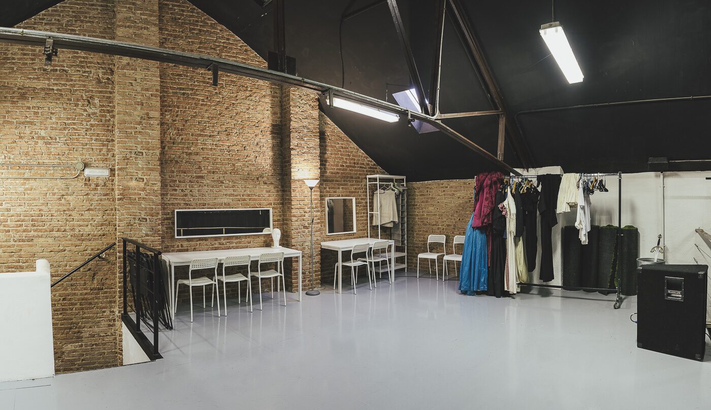 la sala multiusos montada para trabajo: perchas con vestuario, mesas de caballete y sillas frente al muro de ladrillo